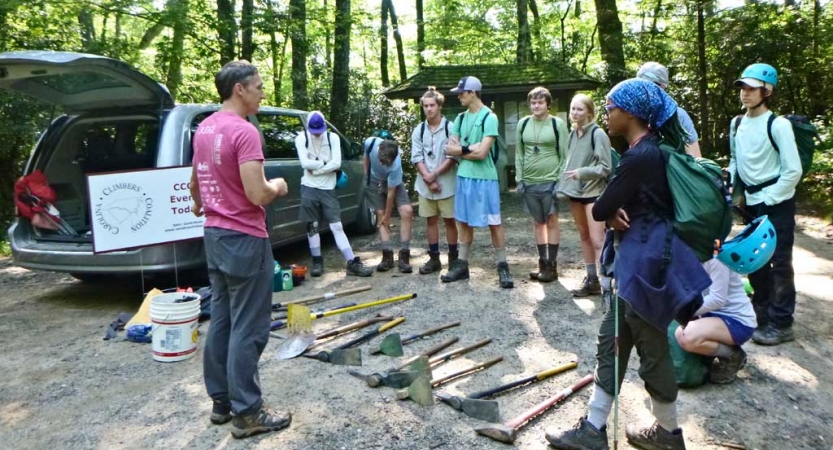 A person speaks to a group of students. There are garden tools laying on the ground between them.